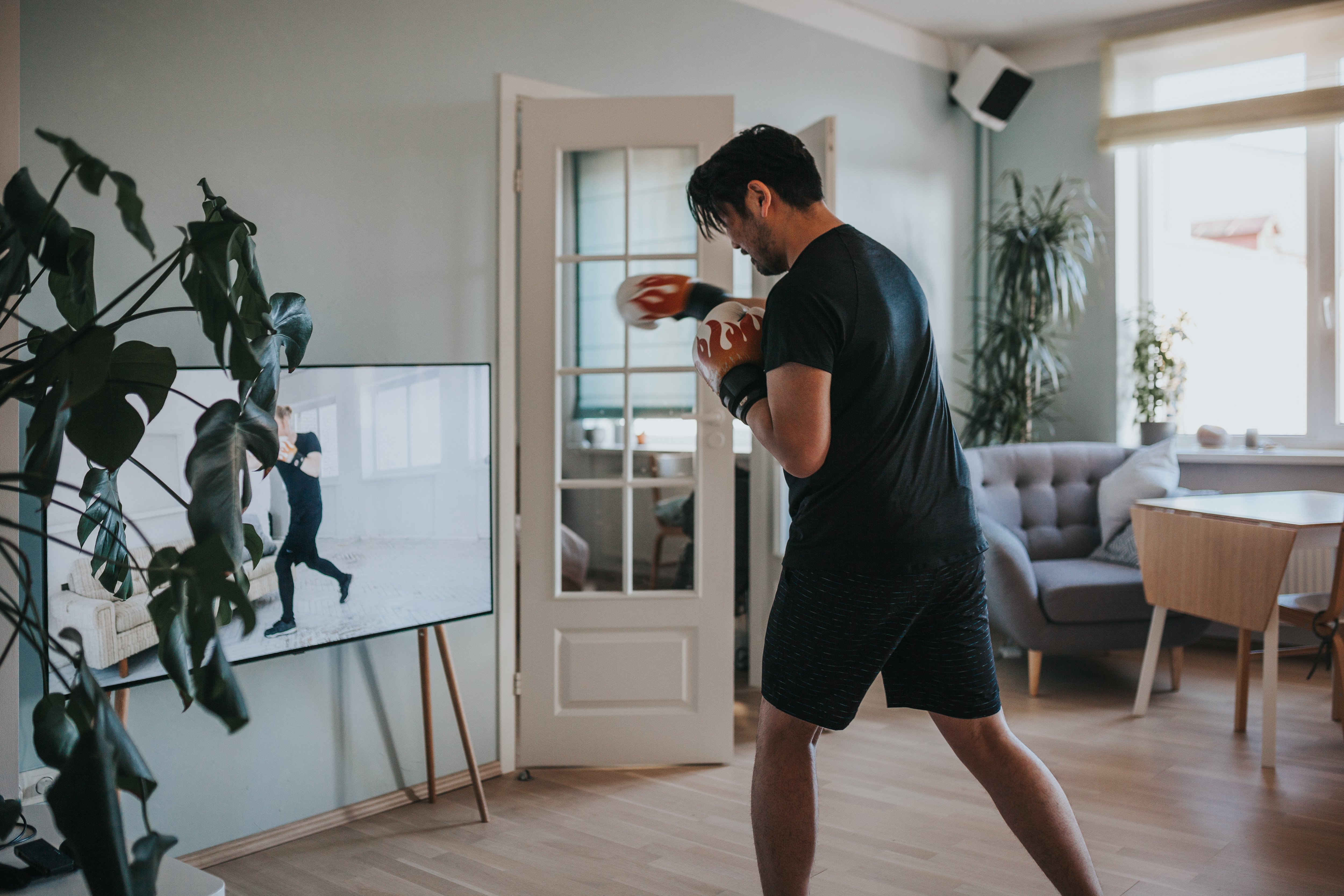 Man in shorts, T-shirt, and boxing gloves doing a kickboxing class with his TV in his den